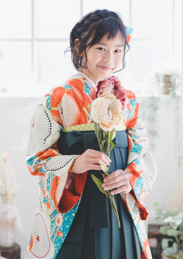 旬色の花柄の着物と深緑色の袴を着た女の子が花を持って微笑んでいるイメージ